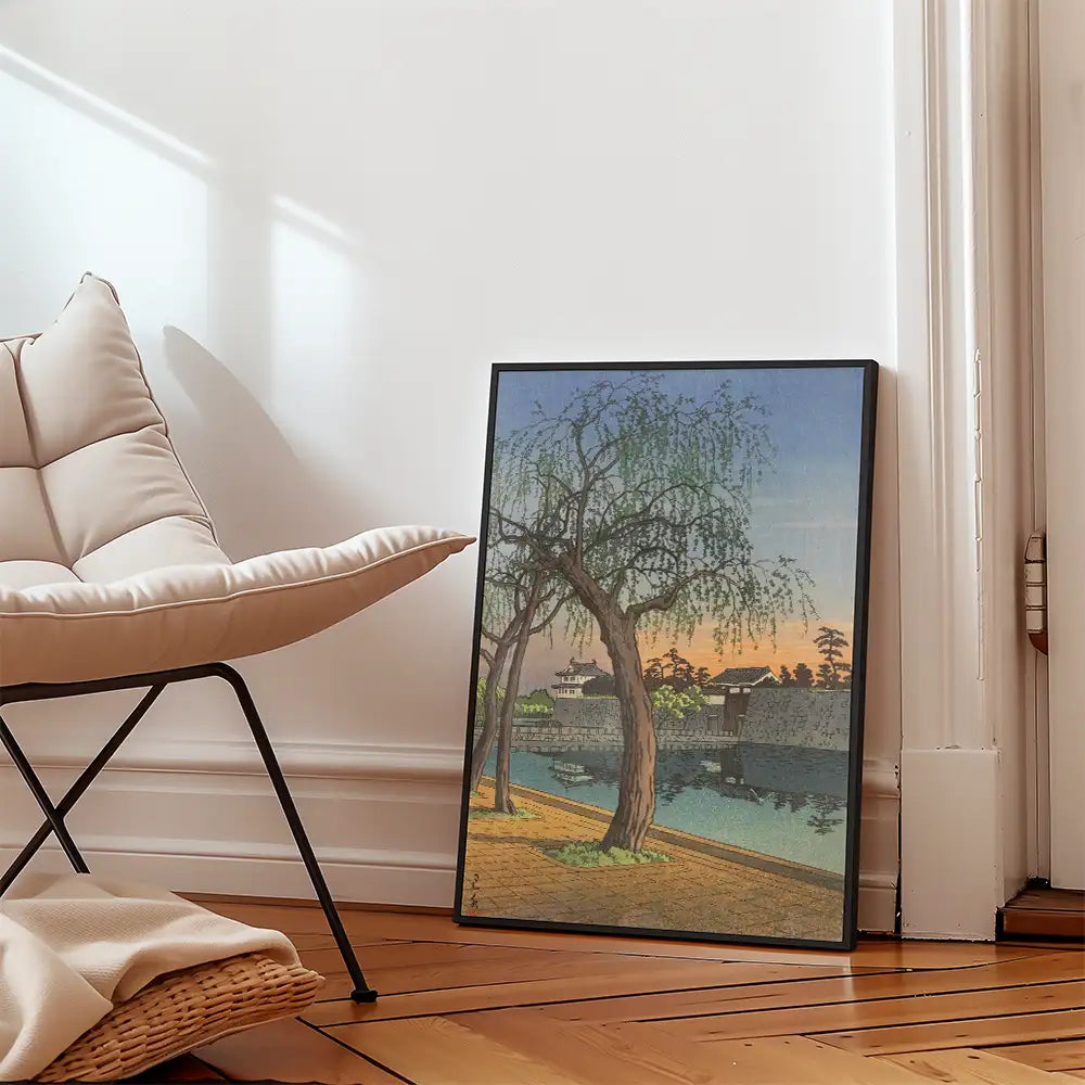Framed painting of a serene landscape with a willow tree by a river, showcased next to a beige chair in a well-lit room.