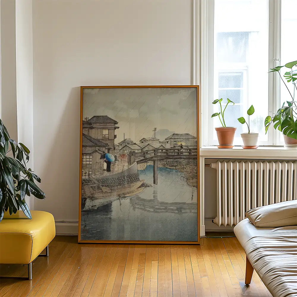Framed painting of a serene riverside village scene featuring a bridge, traditional buildings, and a figure with an umbrella, displayed in a modern living space.