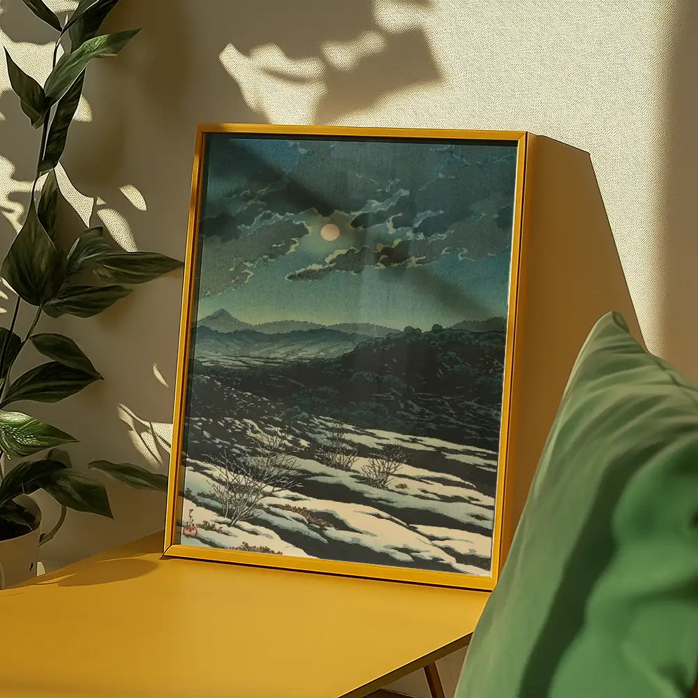 Framed painting of a serene landscape with clouds and a full moon, displayed on a yellow table next to a plant and green cushion.