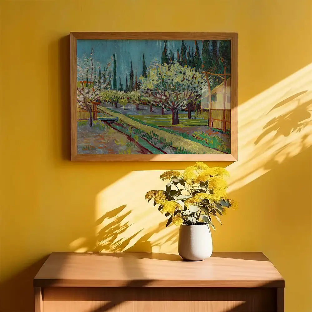 Framed painting of a vibrant landscape with blooming trees, hanging on a yellow wall, alongside a vase of yellow flowers on a wooden surface.