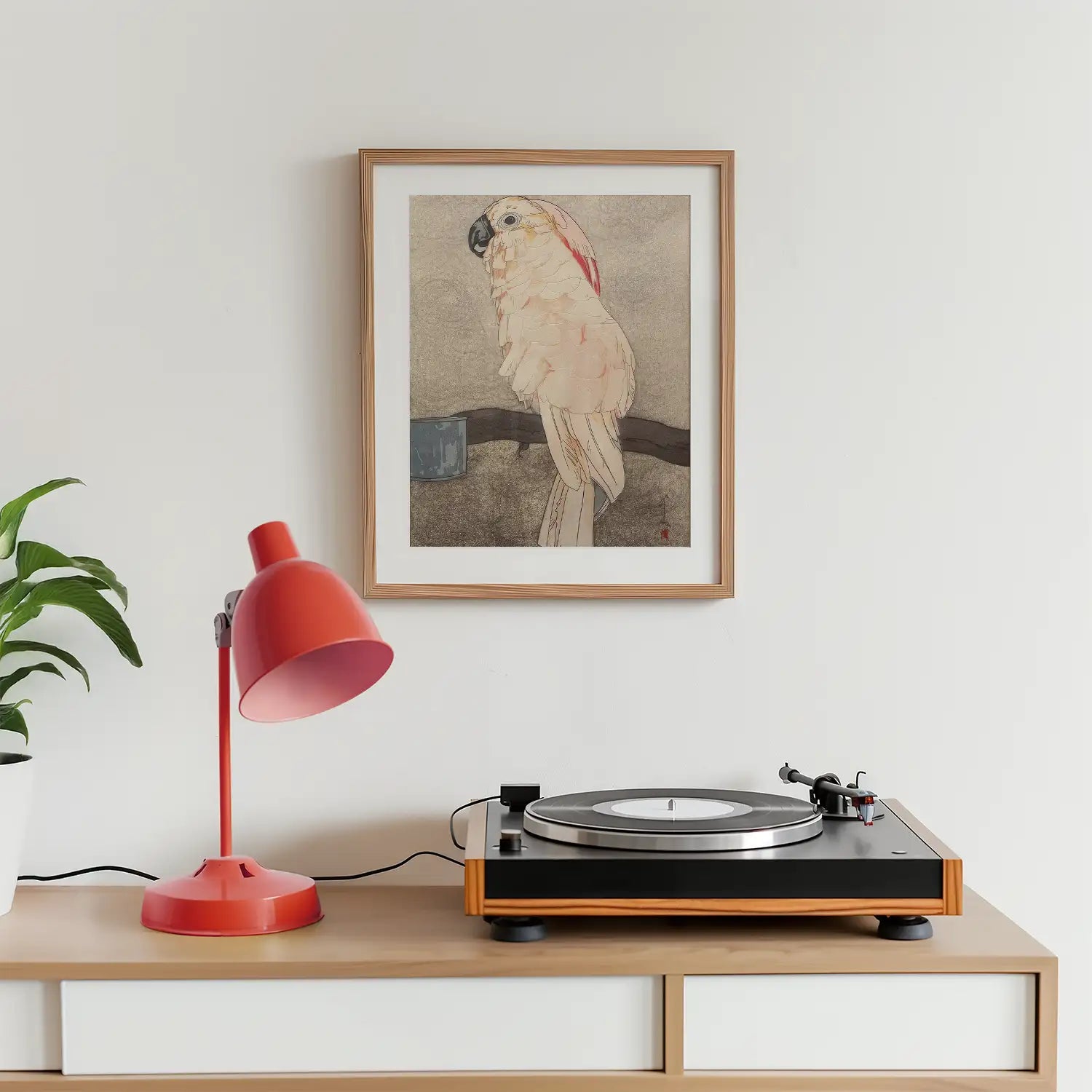 Framed painting of a parrot displayed above a record player and a red table lamp on a wooden surface with a plant to the left.