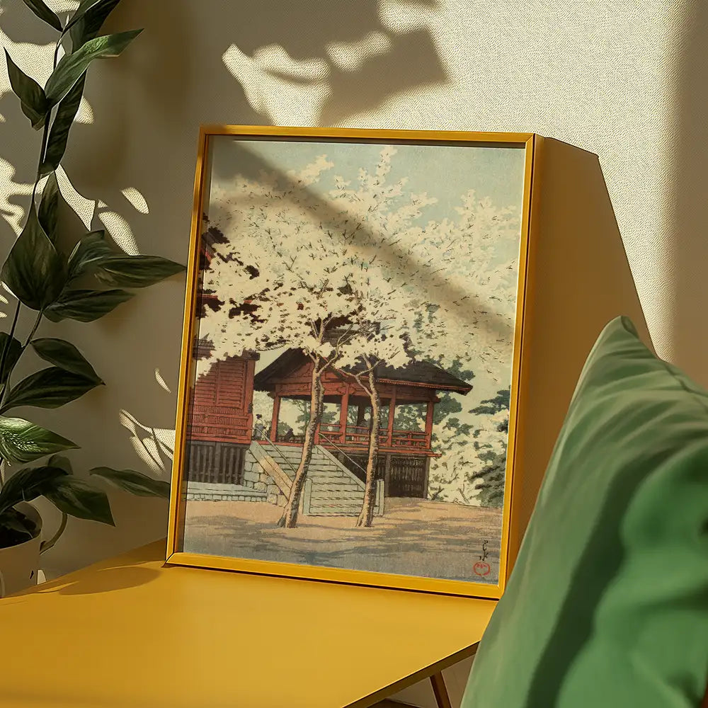 Framed painting of a traditional wooden structure surrounded by cherry blossom trees, placed on a yellow table with a green cushion and indoor plants in the background.
