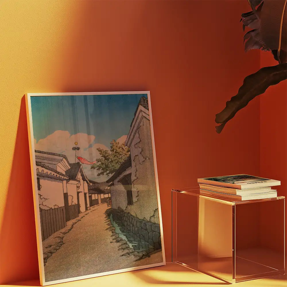 Artistic painting of a serene village pathway, framed and leaning against an orange wall, with a stack of art books on a clear acrylic table.