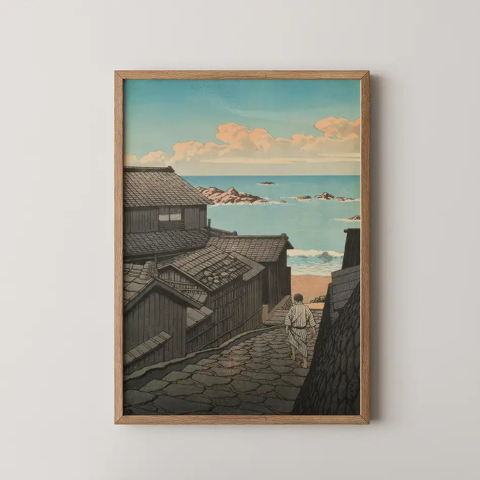 Framed painting of a coastal scene depicting a pathway through traditional buildings leading to the ocean under a blue sky with clouds.