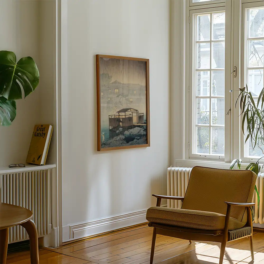 Framed painting of a serene lakeside cabin displayed on a wall in a cozy living room with a plant and a chair.