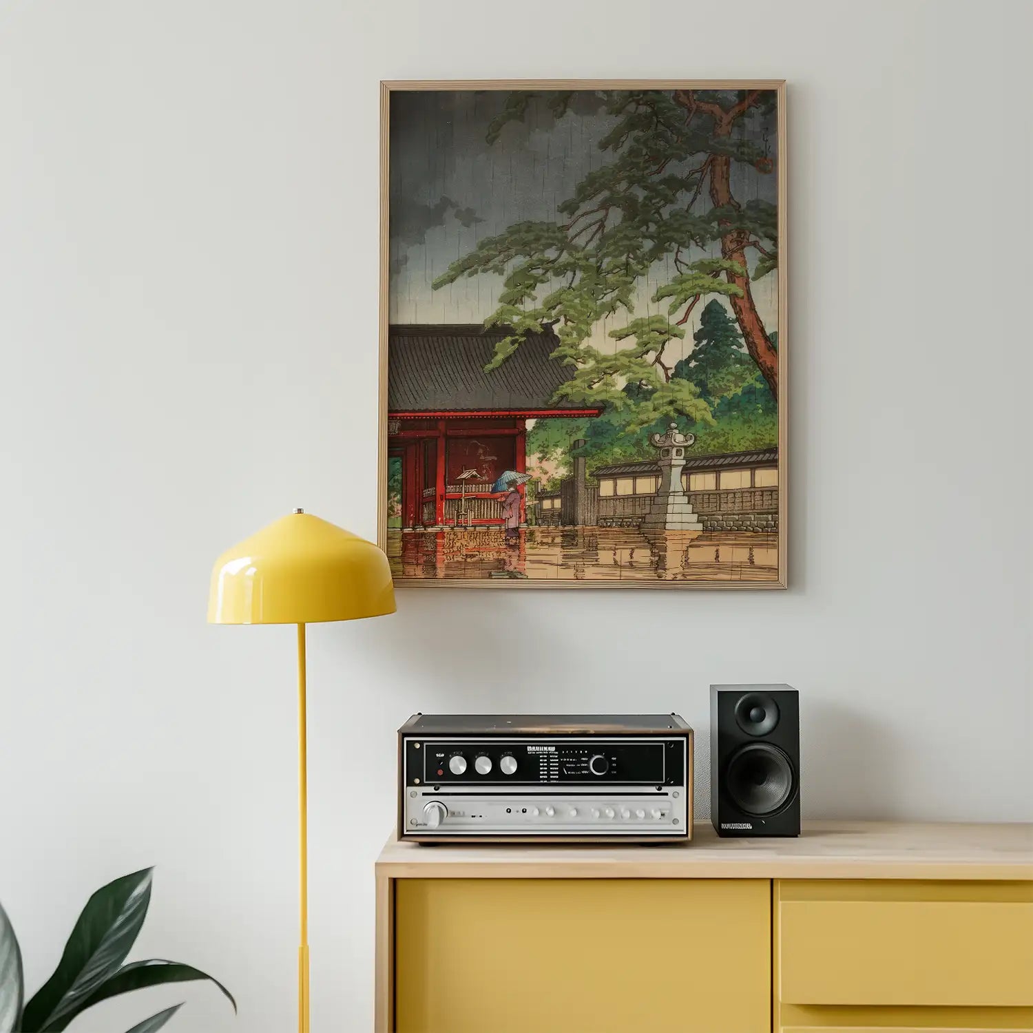 Framed painting of a traditional Asian landscape featuring a red building, surrounded by trees, displayed above a modern record player and yellow lamp in a contemporary living space.