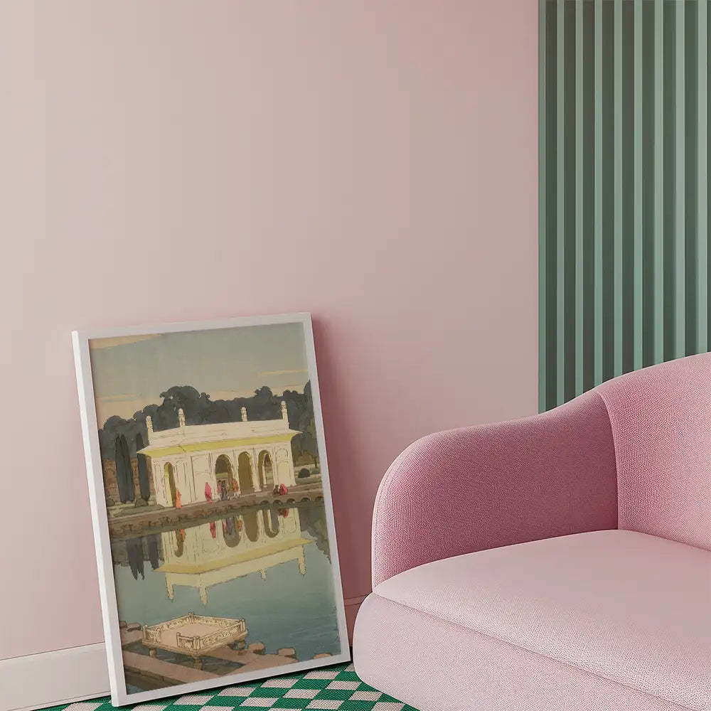 Framed painting of a reflective building by a serene water body, leaning against a pastel pink wall with a pink sofa in the foreground.
