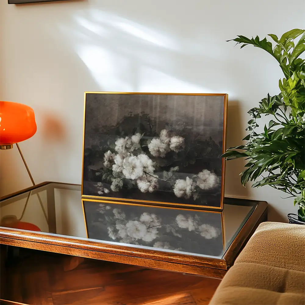 A framed painting of white flowers against a dark background, displayed on a wooden table with a glass top, alongside a green plant and an orange lamp.