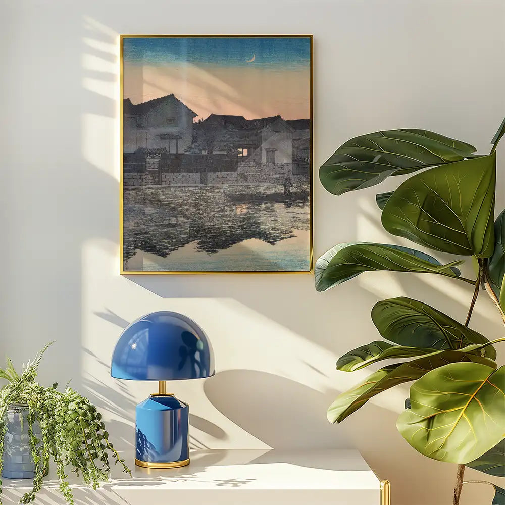 Framed painting of a serene landscape with houses by a water body, displayed on a light wall alongside a blue lamp and green plant.