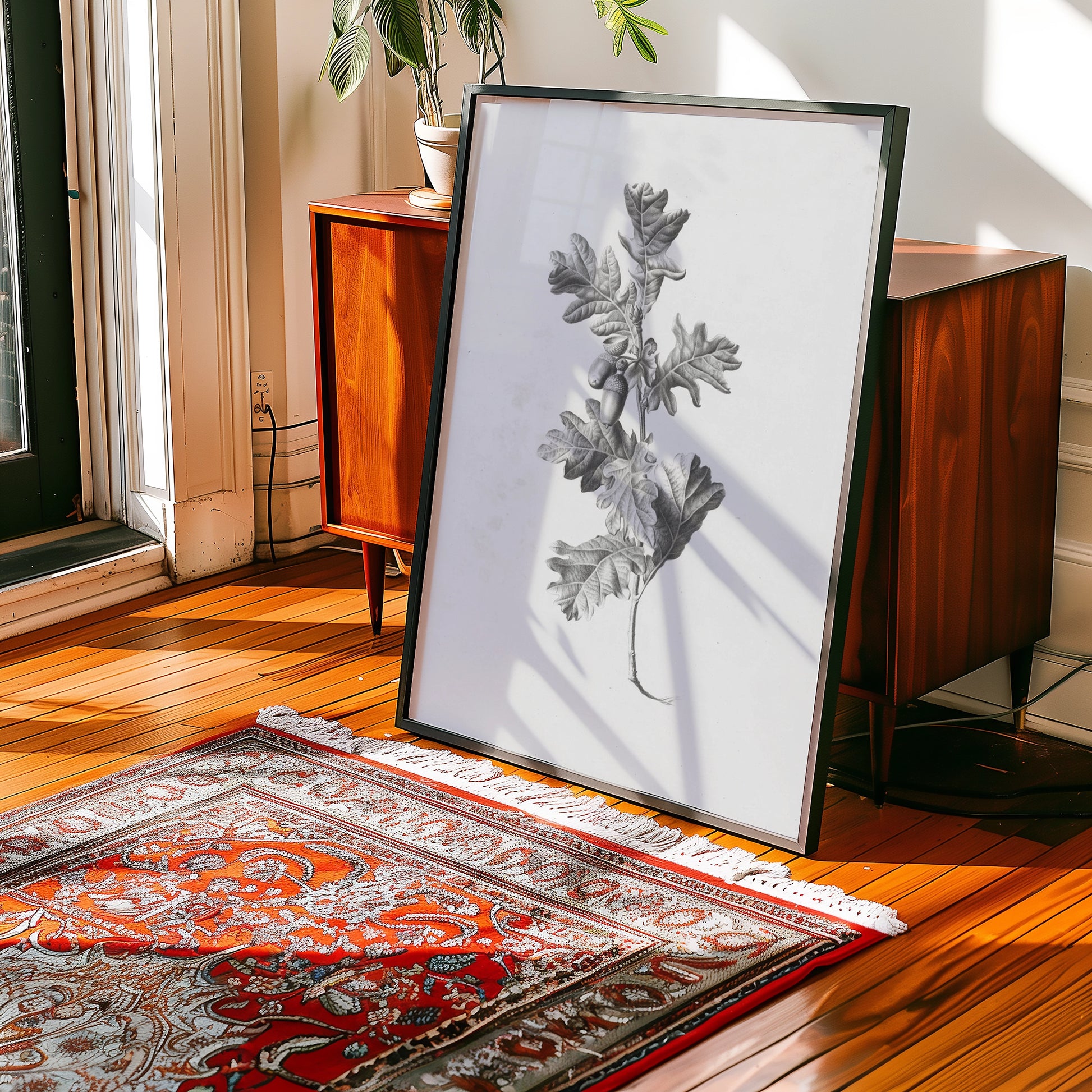 Framed black and white painting of a leaf branch displayed in a sunlit room with wooden flooring and a patterned rug.