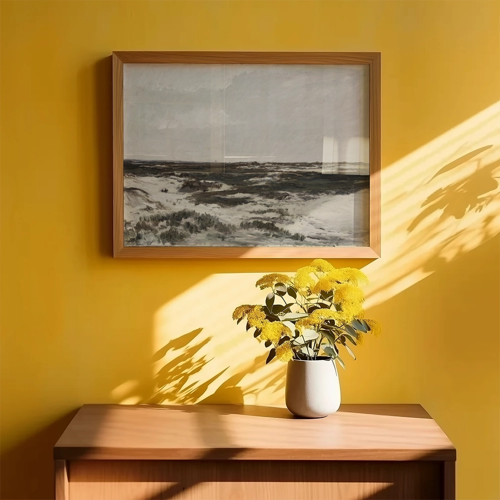 Framed landscape painting on a yellow wall above a wooden table with a white vase filled with yellow flowers.