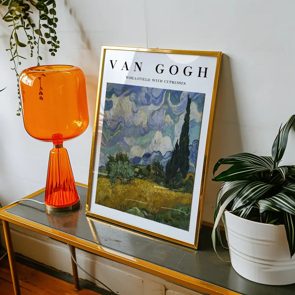 Framed poster of Vincent van Gogh's "Wheatfield with Cypresses" displayed on a table alongside an orange lamp and a potted plant.
