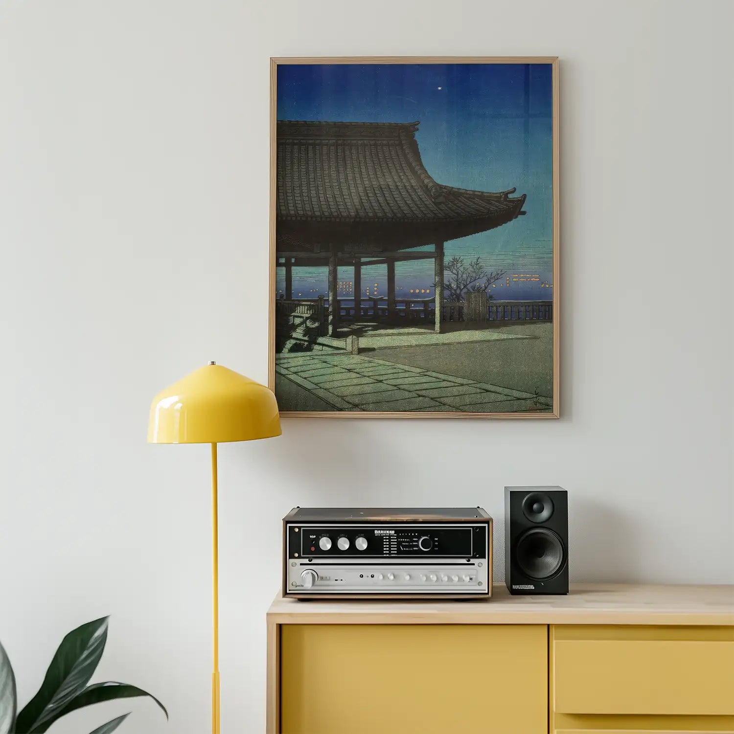Framed painting of a traditional pagoda at dusk, displayed on a white wall above a yellow lamp and a retro audio setup.