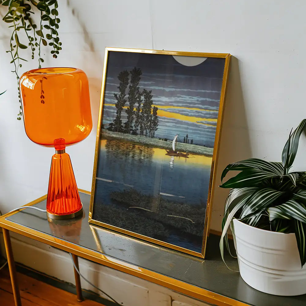 Framed painting of a serene landscape featuring a boat on water at sunset, displayed alongside an orange glass lamp and a potted plant.
