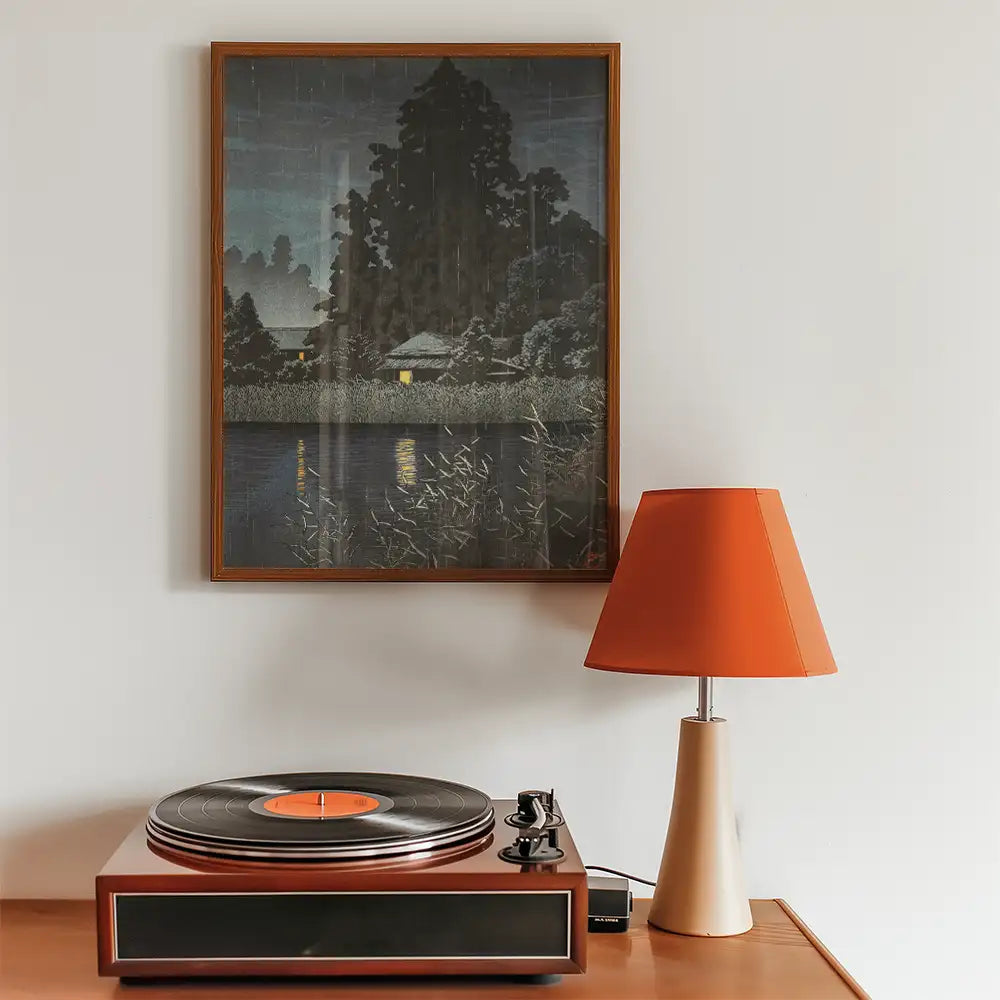 Painting of a serene night scene with a house by a reflective pond, framed and displayed above a vintage record player and an orange lamp.