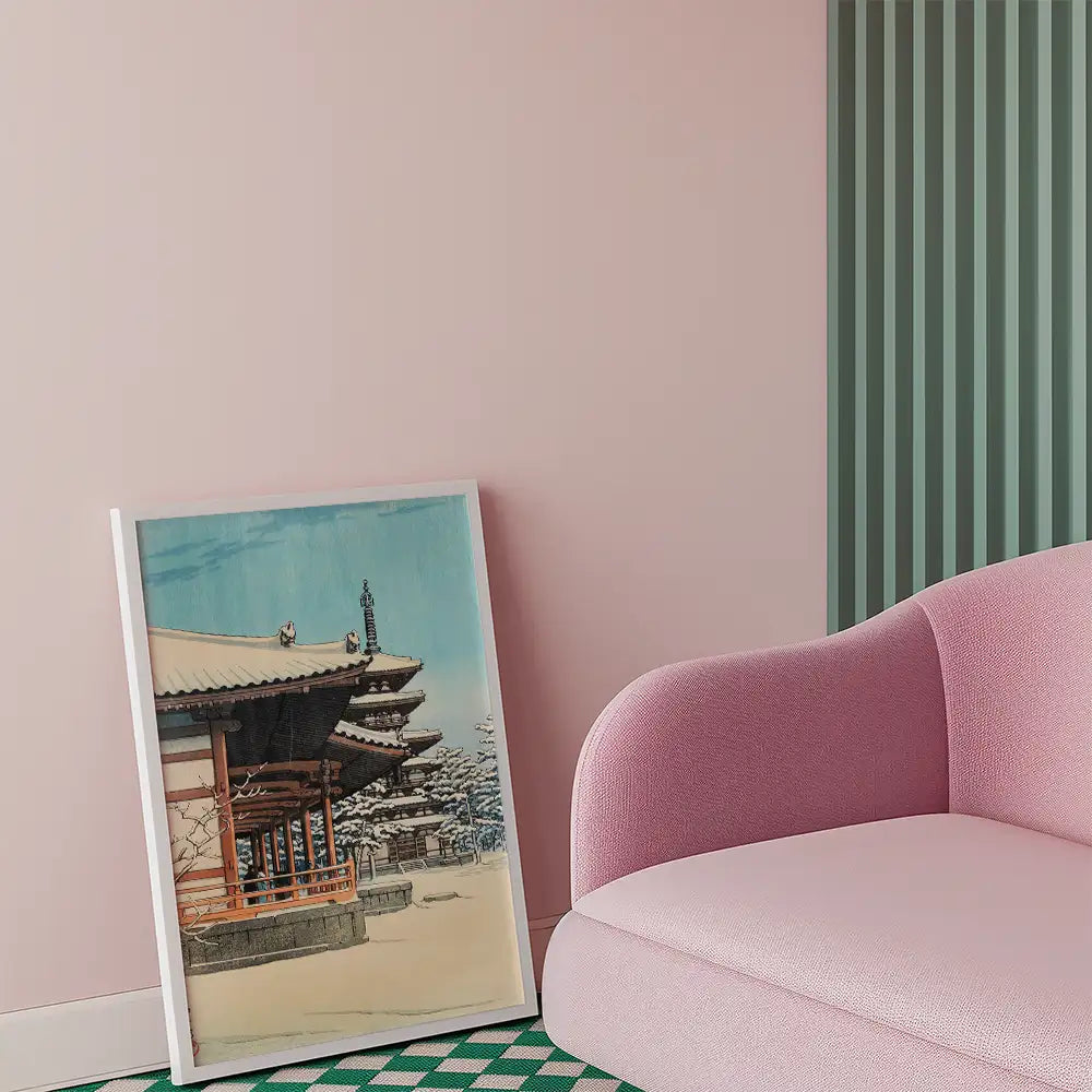 Framed painting of a traditional building with a snow-covered landscape, leaning against a pink wall next to a soft pink sofa.