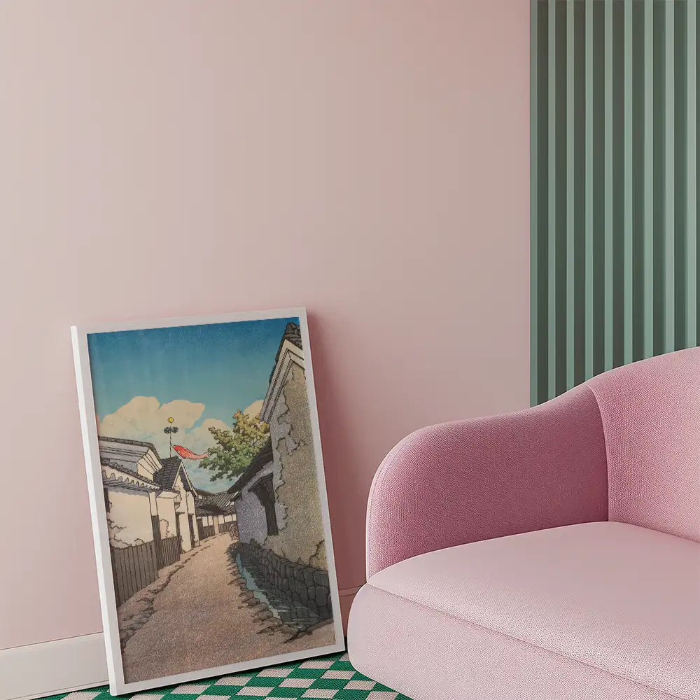 Framed painting of a serene street scene with traditional houses and a blue sky, leaning against a pink sofa in a stylish interior setting.