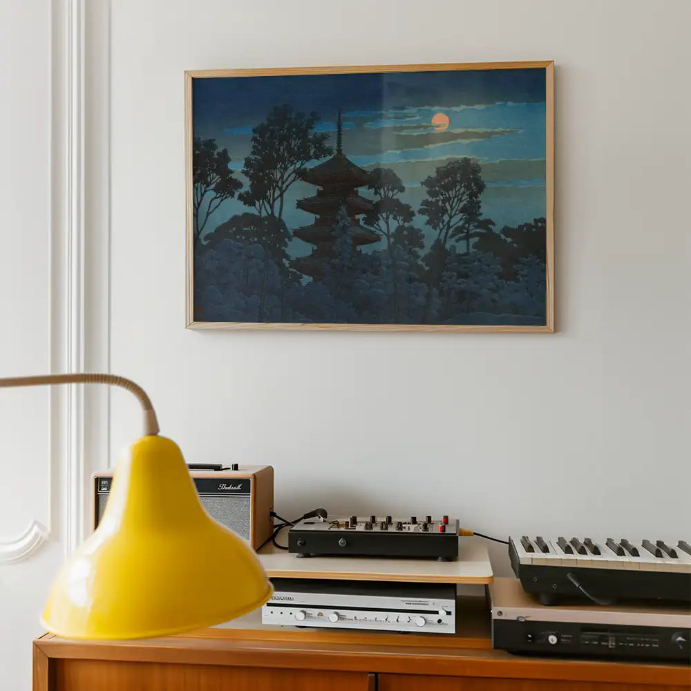 Framed painting of a pagoda against a night sky with trees, hanging on a white wall above a wooden console table featuring audio equipment and a yellow lamp.