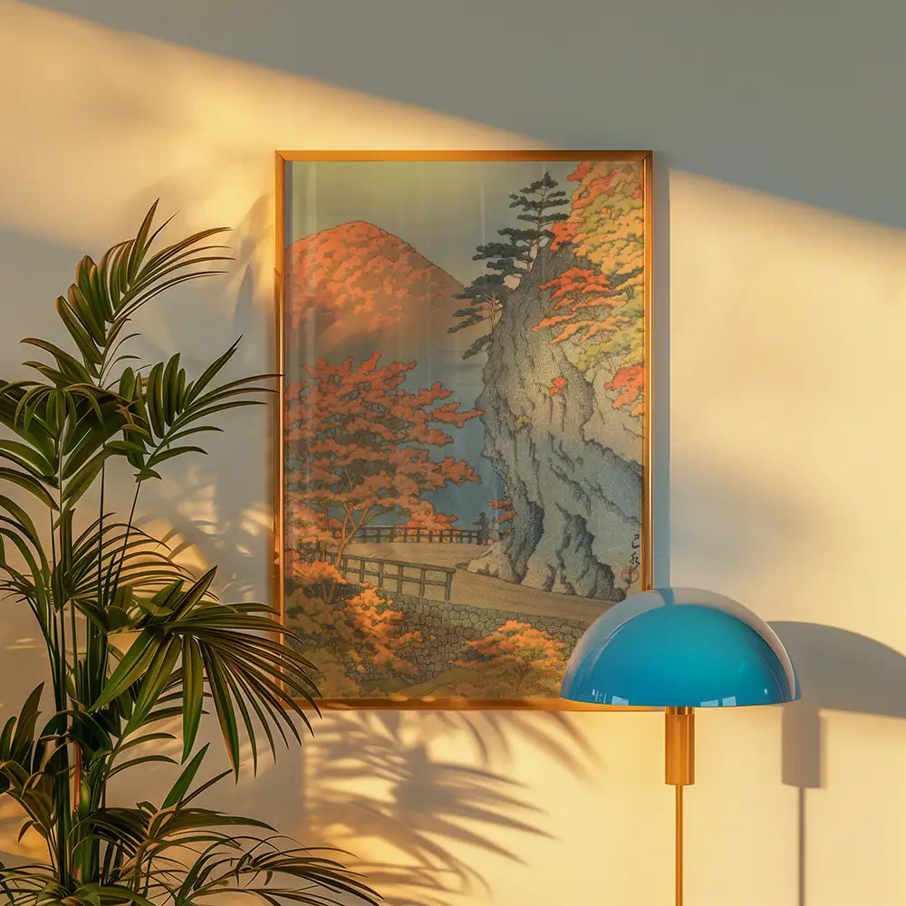 Framed painting of a scenic landscape featuring autumn foliage, mountains, and a pathway, displayed on a wall next to a green plant and a blue lamp.