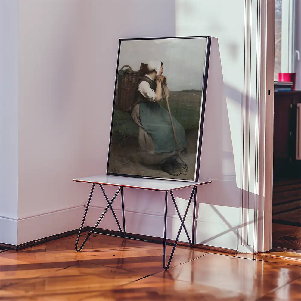 A framed painting of a woman sitting with a basket, displayed on a minimalist table in a bright room.