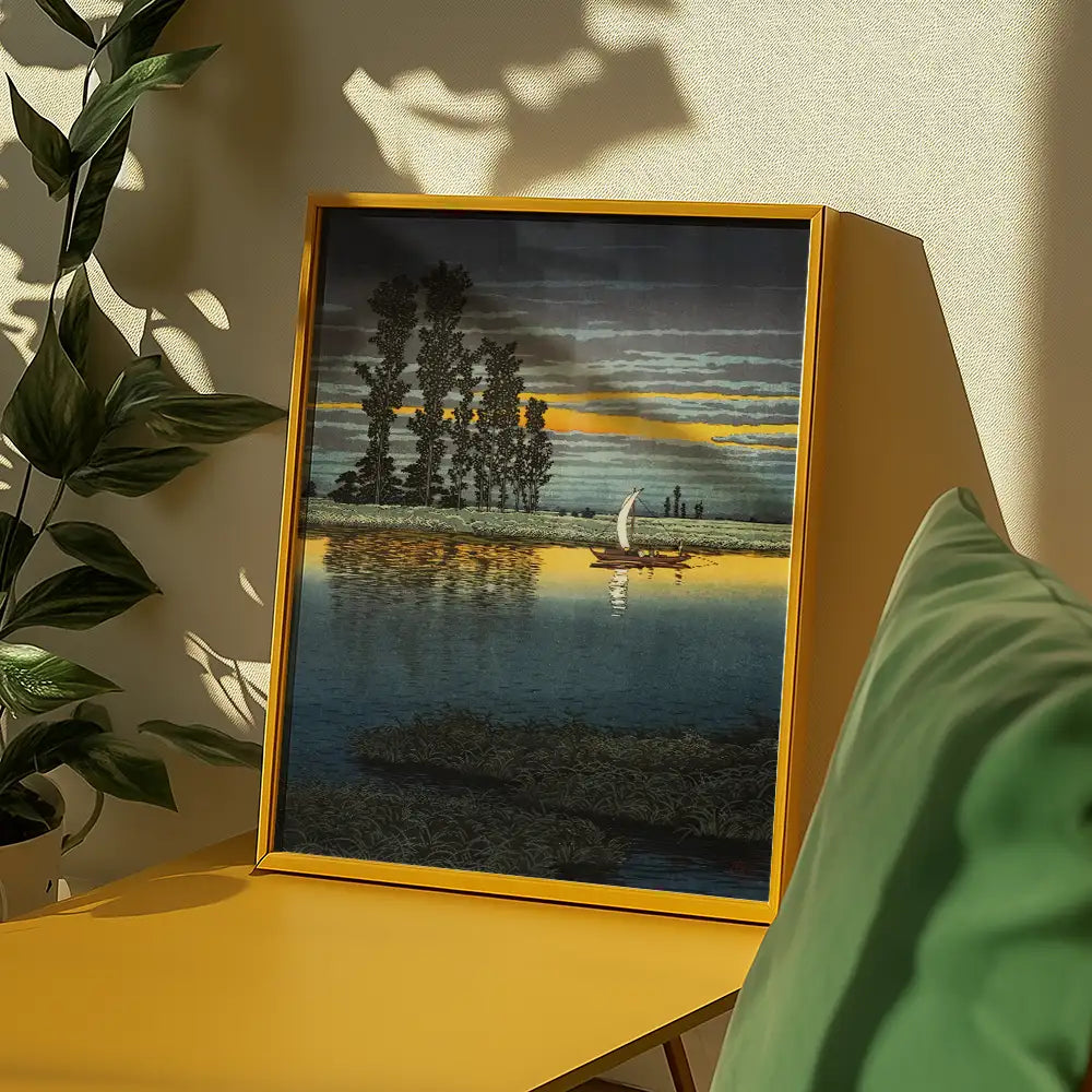 Framed painting of a serene lakeside landscape at sunset, featuring a small sailboat on calm waters surrounded by tall trees and vivid clouds, displayed on a yellow table next to green cushions.