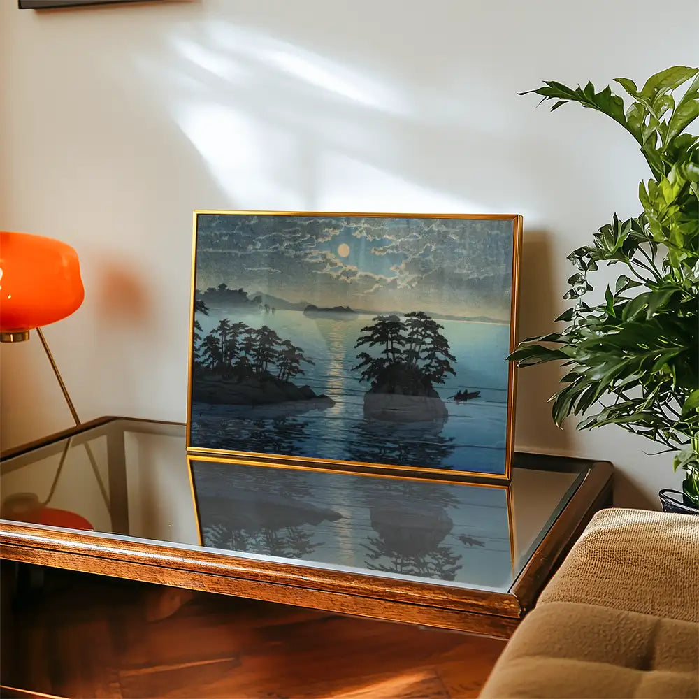 Framed painting of a serene landscape featuring trees and mountains reflecting in water under a cloudy sky, placed on a wooden table with a glass surface and a decorative plant nearby.