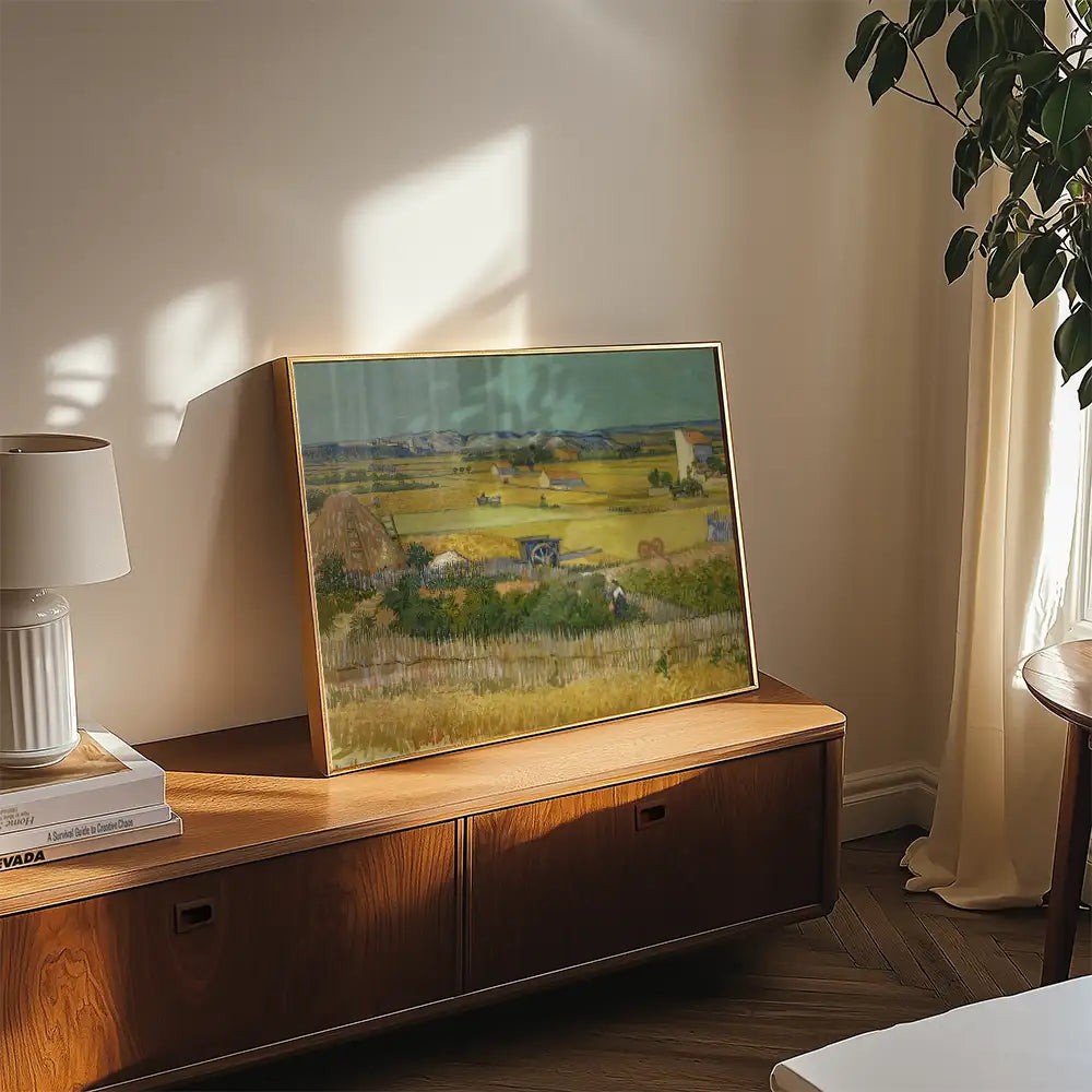 Painting of a serene countryside landscape featuring fields, haystacks, and distant buildings, displayed on a wooden side table with a lamp and potted plant in a well-lit room.