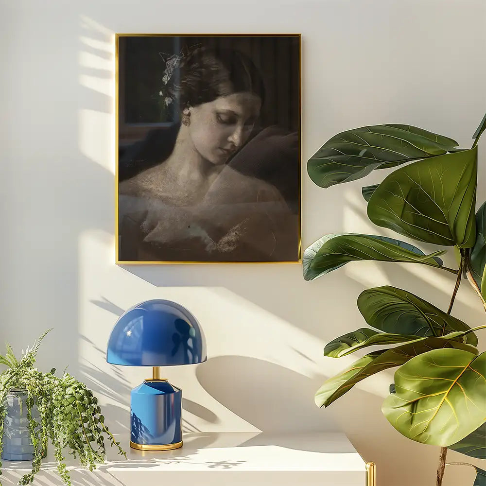 Portrait painting of a woman in a gold frame, displayed on a wall above a stylish blue lamp and green plant on a white table.