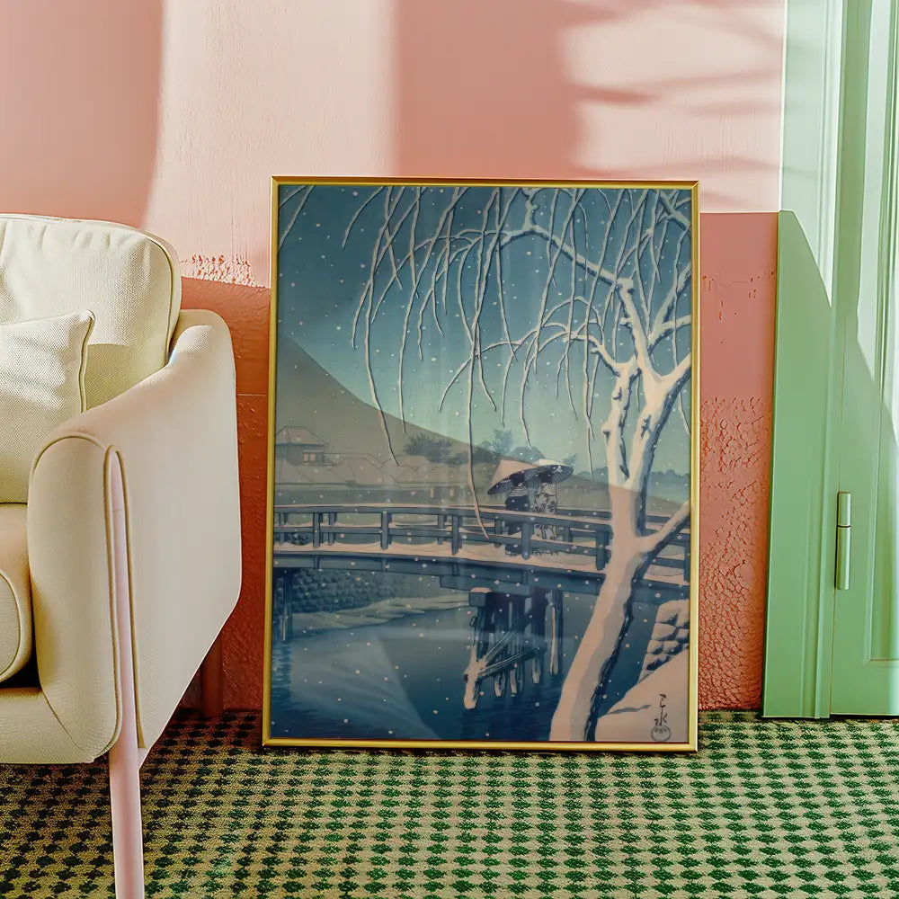 A framed winter landscape painting featuring a serene snowy scene with a bridge and a figure under an umbrella, placed beside a light-colored armchair in a stylish room.