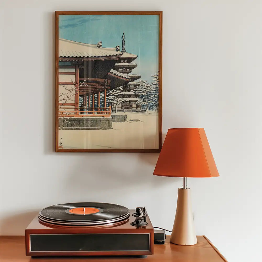Framed painting of a traditional Japanese building in winter, displayed above a wooden record player and a modern orange lamp.