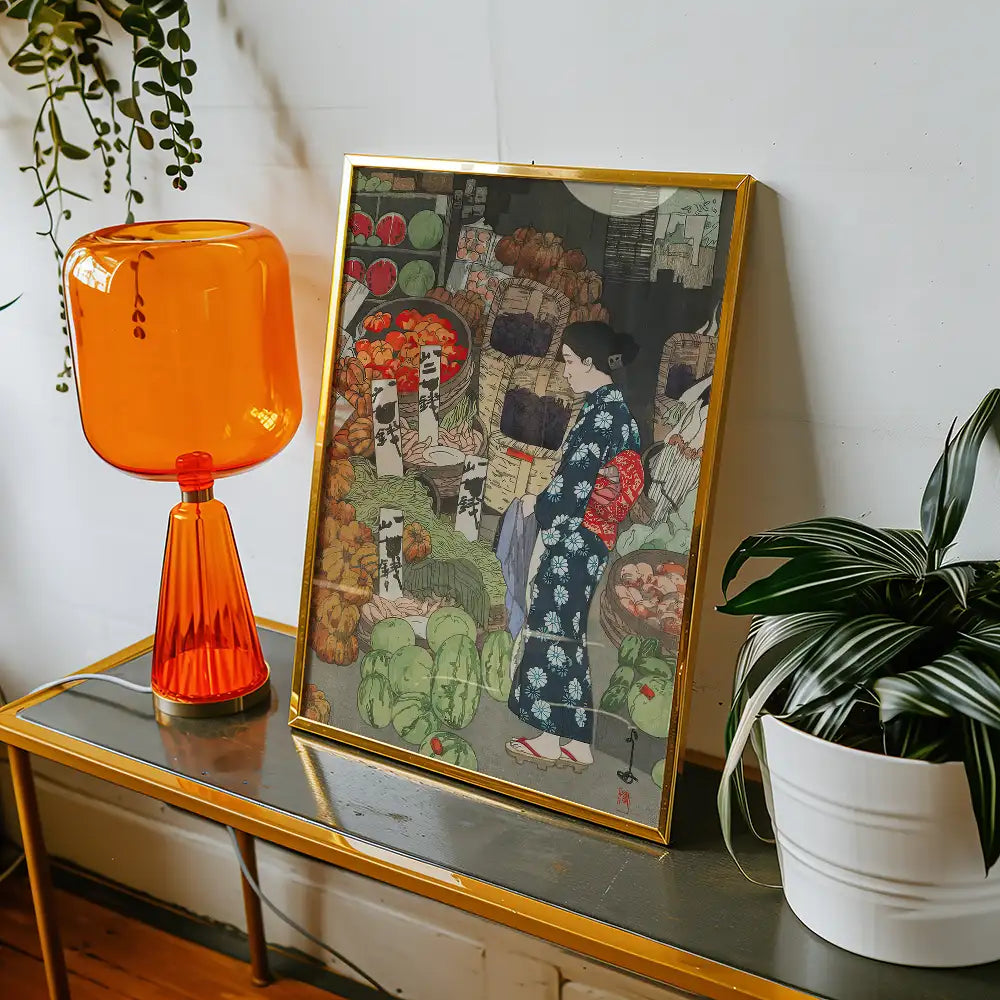 Painting of a woman in a blue kimono standing at a vibrant market with fruits and vegetables, framed and displayed on a side table next to an orange lamp and a potted plant.