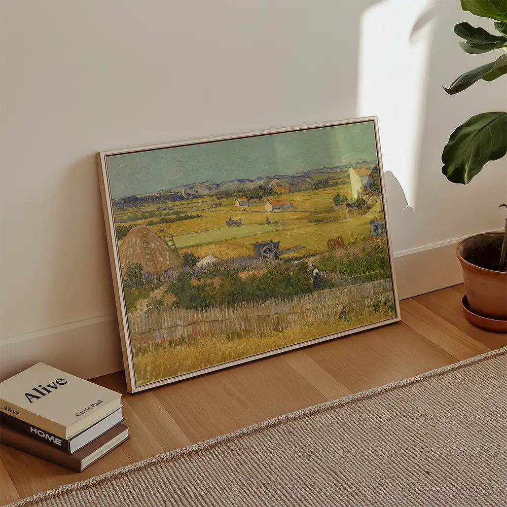 Framed painting of a pastoral landscape with fields, farmhouses, and distant mountains, leaning against a wall with books and a plant nearby.