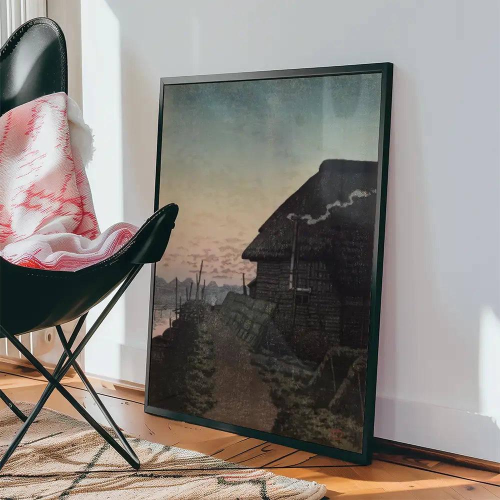 Framed painting of a rustic cottage at sunset, leaning against a wall beside a chair with a patterned throw blanket.