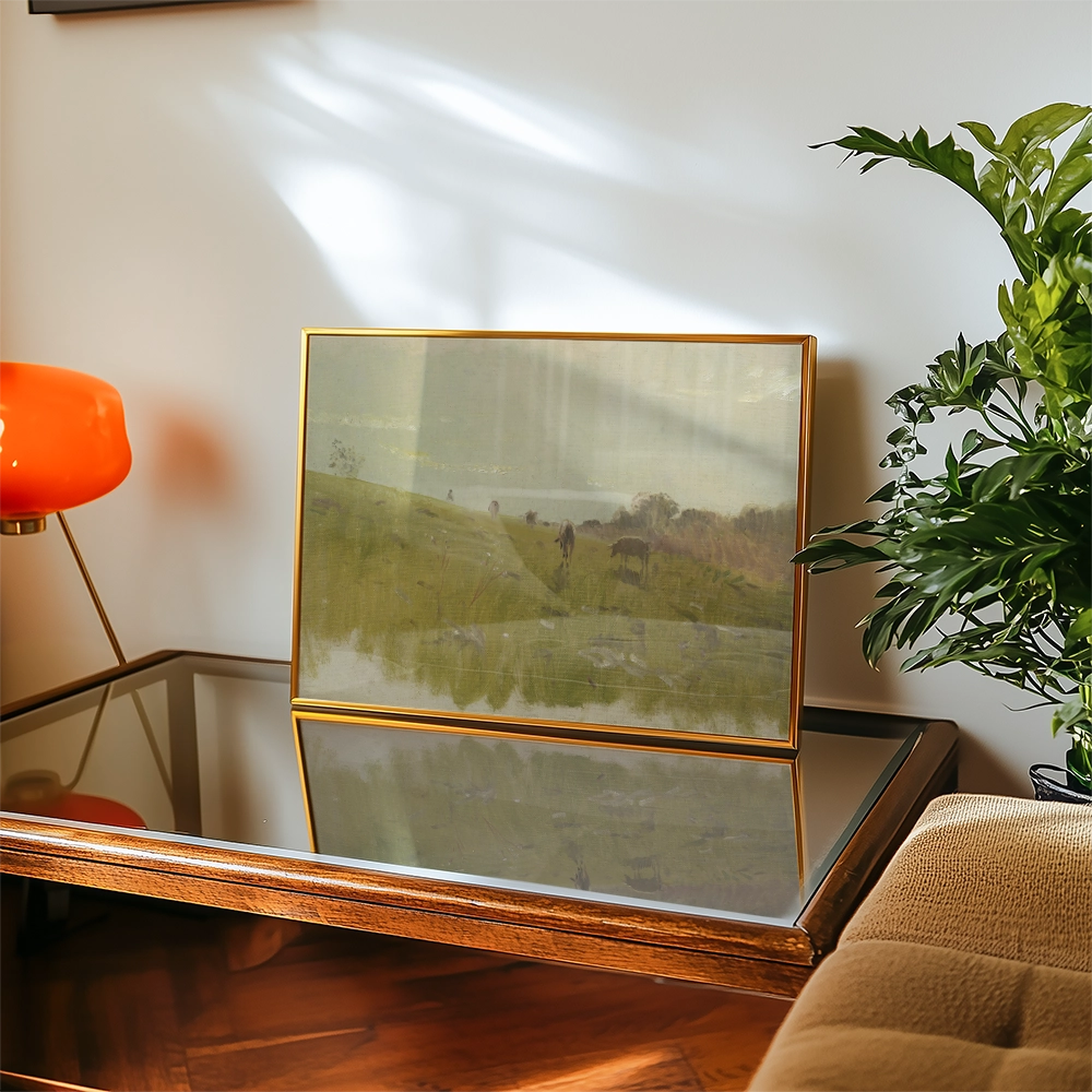 Framed landscape painting displayed on a glass table with a plant and a lamp in a cozy interior setting.
