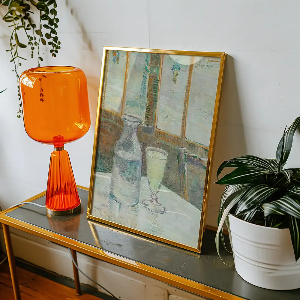 Framed painting of a glass and a bottle on a table, accompanied by an orange lamp and a potted plant.