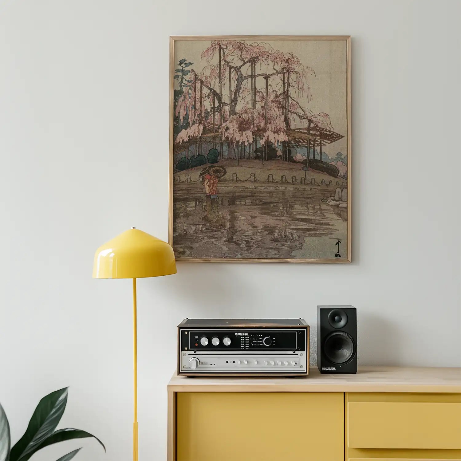 Framed painting of cherry blossom trees by a pond, displayed above a yellow lamp and a vintage audio system on a minimalist wooden cabinet.