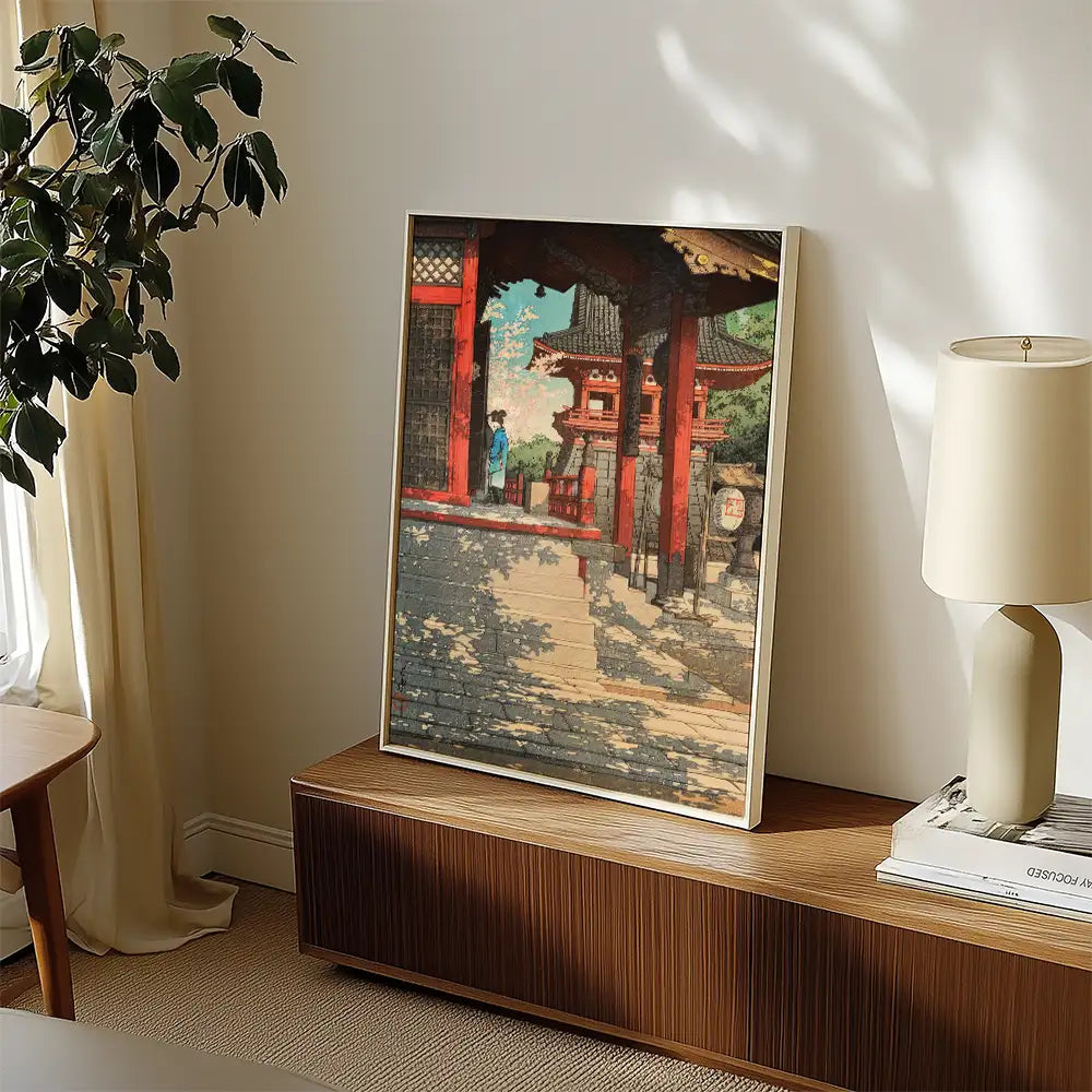 Framed painting of a traditional temple setting with a figure standing on a pathway, displayed on a wooden console table in a sunlit room.