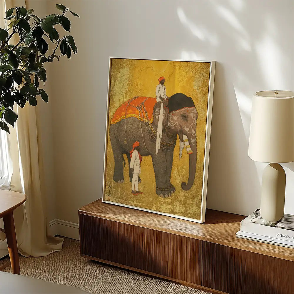 Framed painting of an elephant with two men in traditional attire, displayed on a wooden surface next to a lamp and decorative plant.