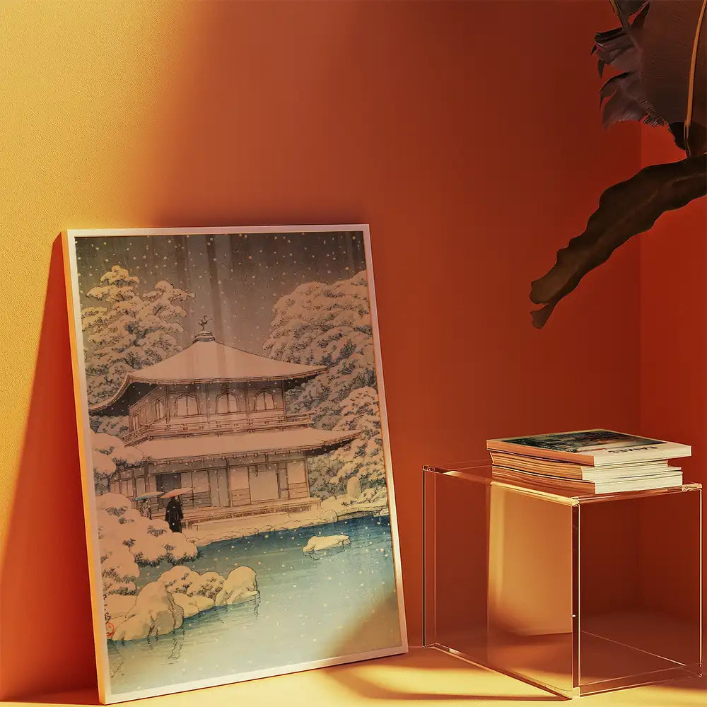 Framed painting of a traditional Japanese temple surrounded by snow and trees, placed against an orange wall with a clear acrylic table holding a stack of books.