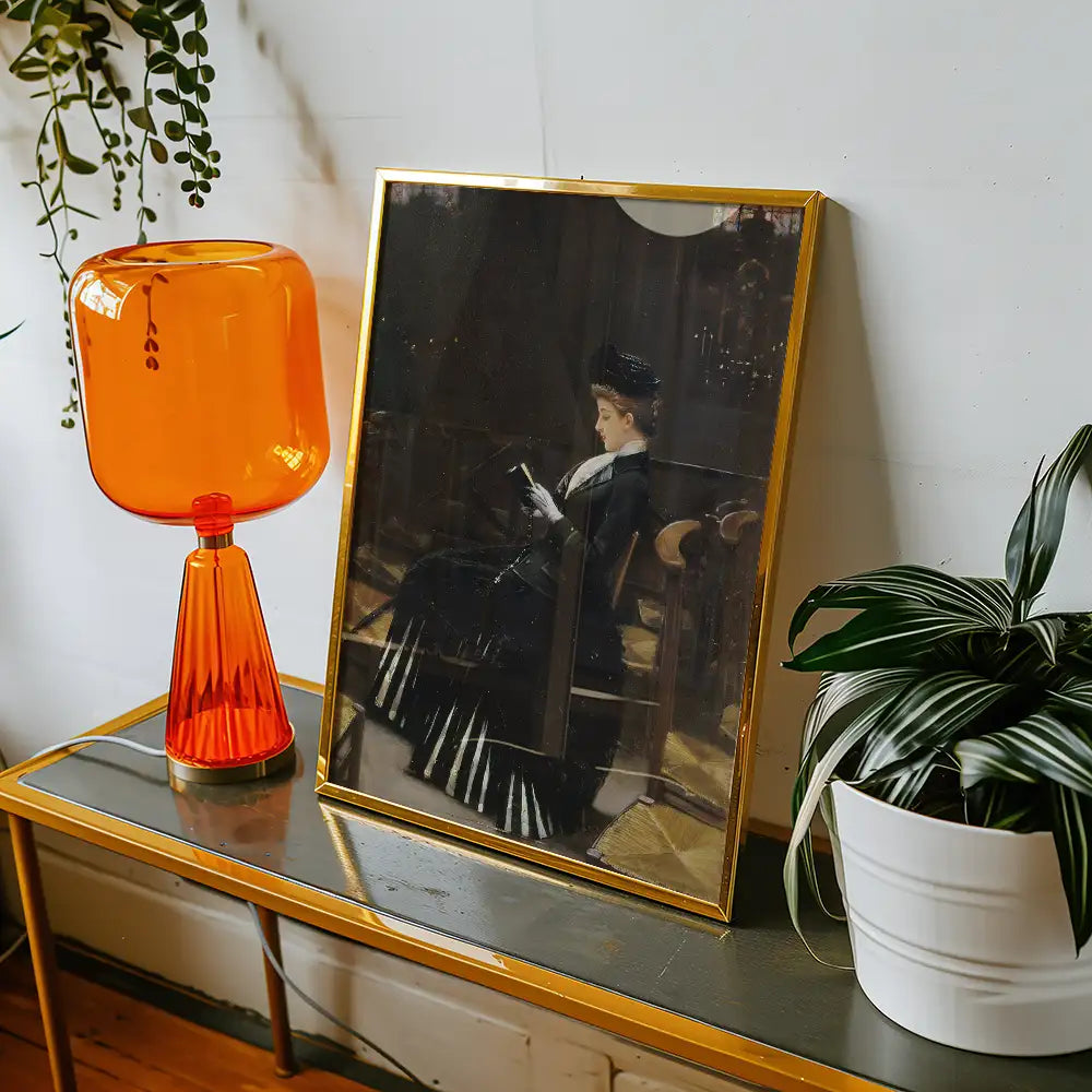 A vintage painting of a woman reading, displayed in a gold frame on a table with an orange lamp and a green plant.