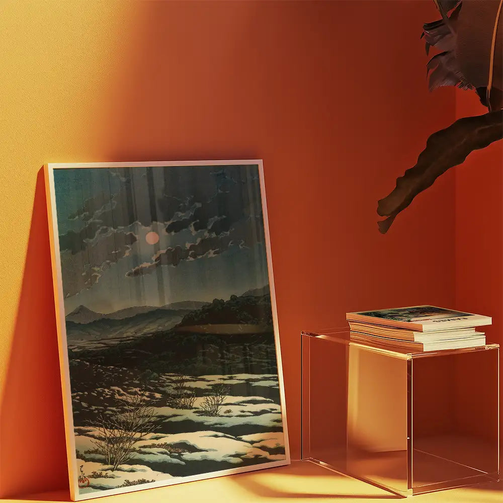 Framed landscape painting featuring mountains and a sunset, displayed against an orange wall with a clear acrylic side table and stacked art prints.