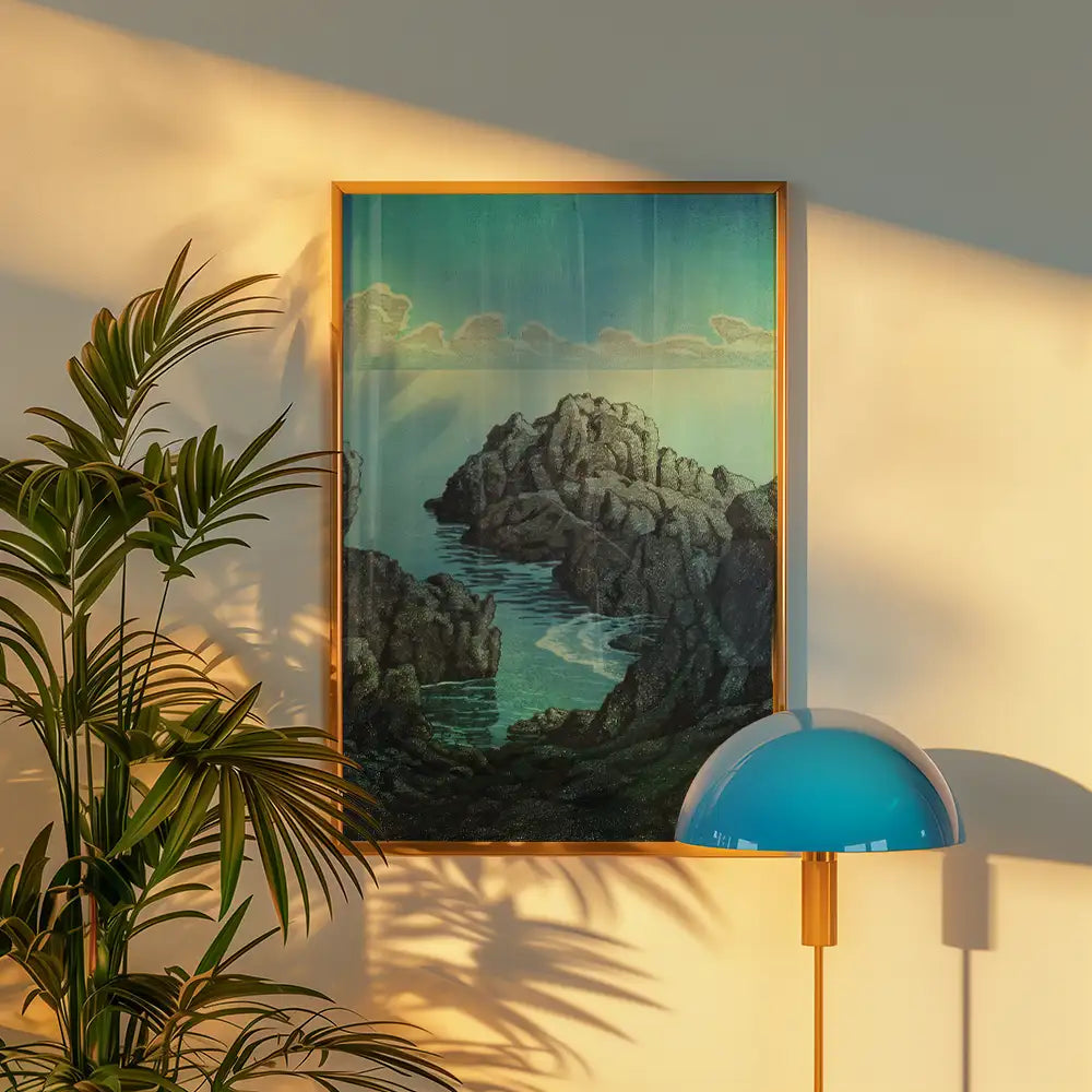 Painting of rocky coastline with turquoise water, framed in wood, displayed on a wall with a green plant and blue lamp in the foreground.