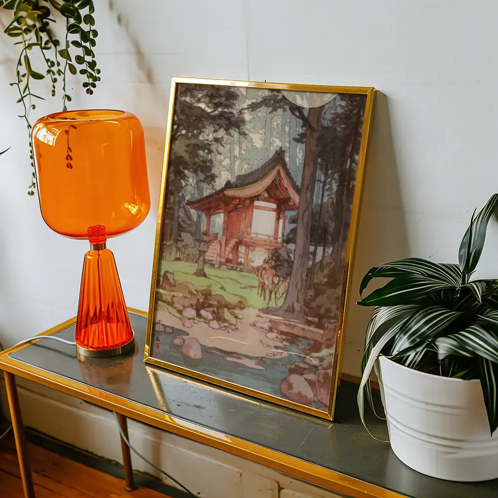 Framed painting of a traditional building surrounded by trees, displayed on a side table next to an orange lamp and a potted plant.