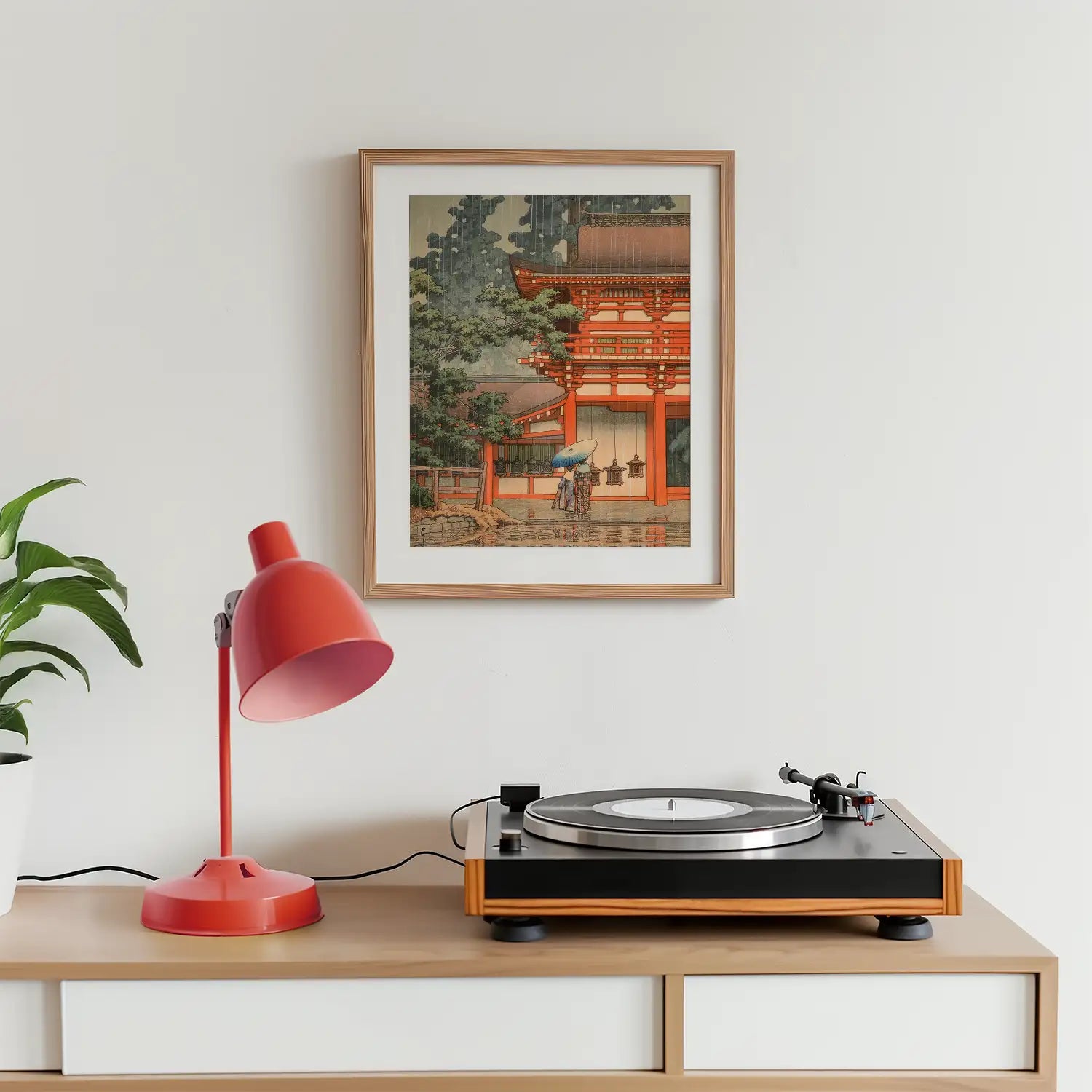 Framed painting of a traditional Japanese architecture displayed on a wall above a wooden console table with a red desk lamp and a turntable.