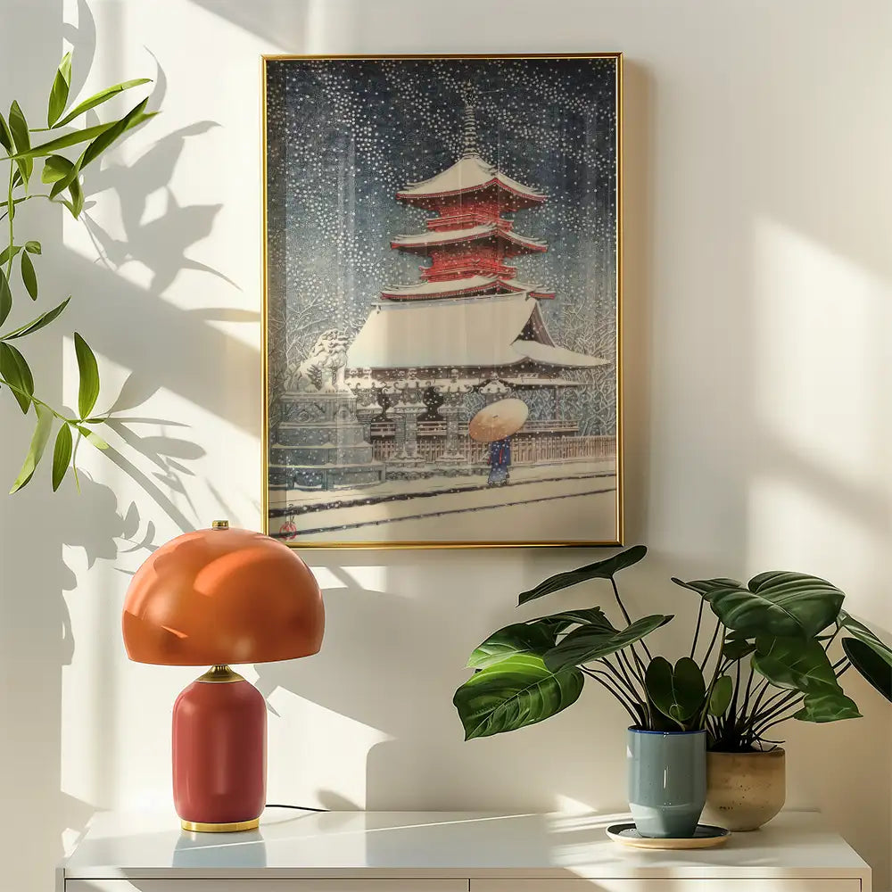 Painting of a traditional Japanese pagoda in a snowy landscape, framed and hung on a wall next to a stylish table lamp and potted plants.