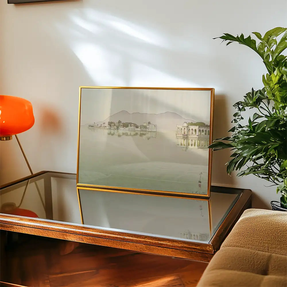 Framed painting of a serene landscape featuring water and mountains displayed on a wooden table with a glass surface and a green plant.