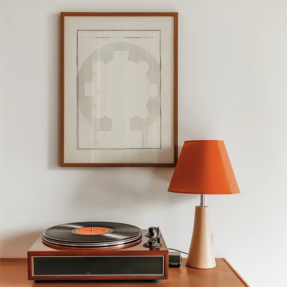Abstract framed wall art above a wooden record player and an orange lampshade, set against a light-colored wall.