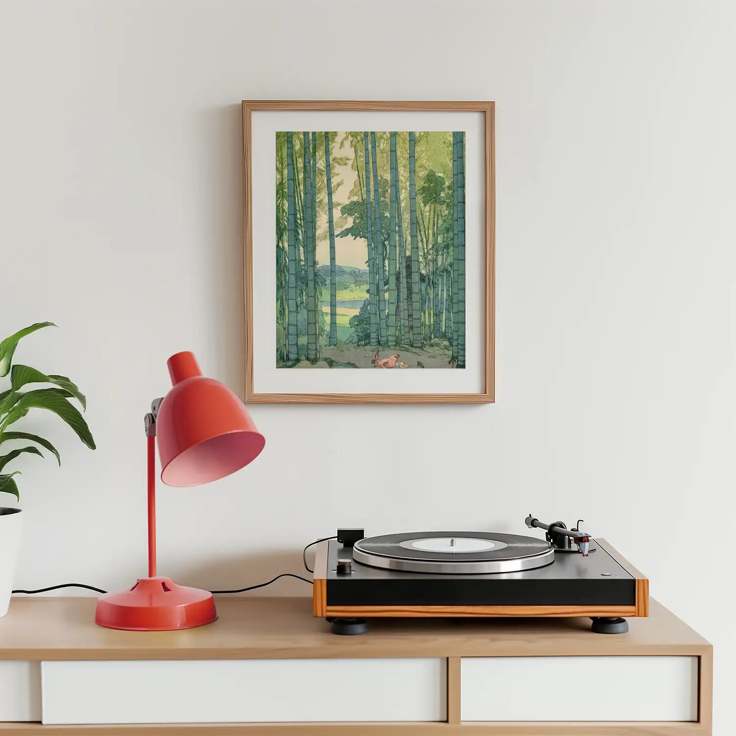 A framed painting of a bamboo forest hanging on a wall above a wooden side table with a red desk lamp and a record player.