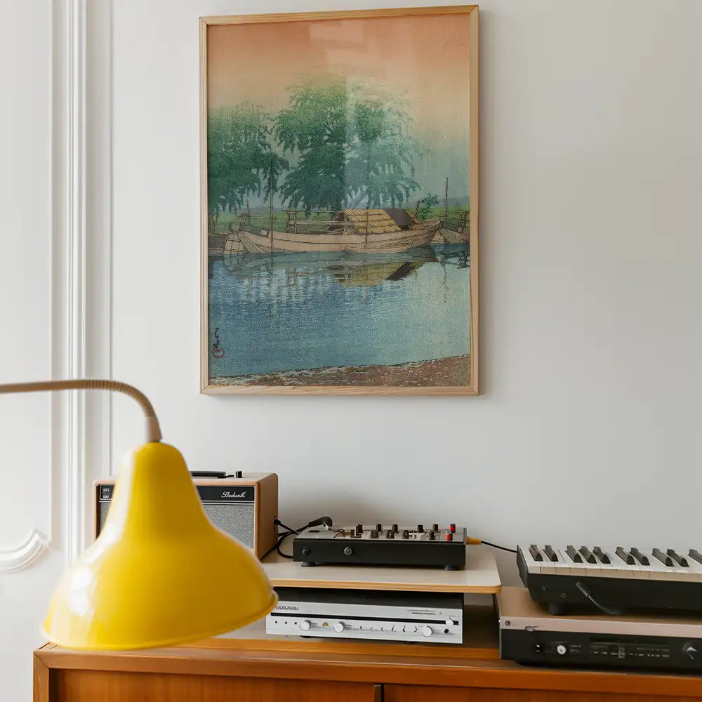 Framed painting of boats on a calm river with trees in the background, displayed on a wall above a wooden furniture piece with a yellow lamp and audio equipment.
