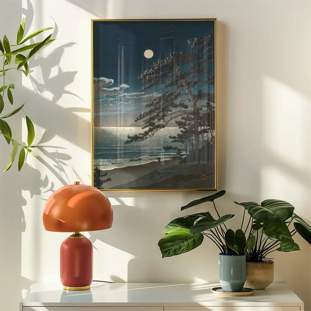 Framed painting of a moonlit beach scene with trees, displayed on a wall alongside a stylish lamp and potted plants.